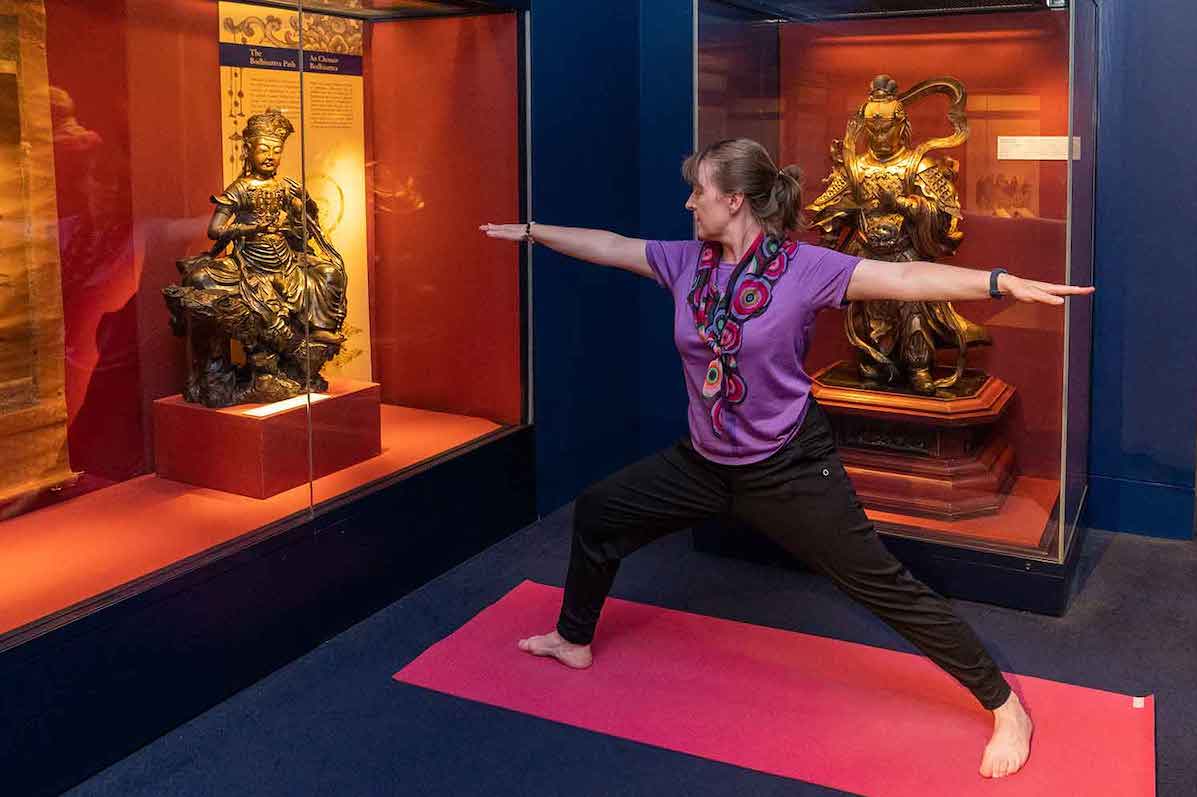 A person in yoga pose with arms outstretched in museum with large display cabinets containing gold figures.