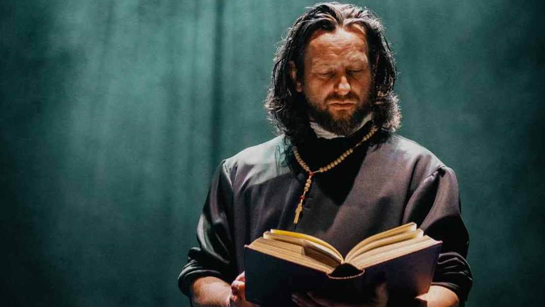 A man with dark shoulder length hair, in a dark top with a wooden rosary around his neck, holding a large, thick old style book, appearing to be reading it, against a dark green background with a light shining down from above.