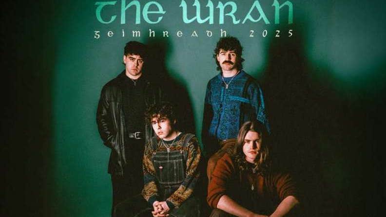 Group of 4 men, 2 standing behind 2 seated on small white blocks, against dark green background.