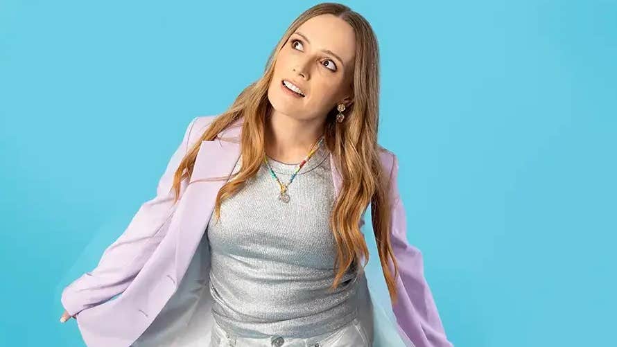 A woman is looking with interest up to her right, holding out either side of a lilac jacket against blue background.