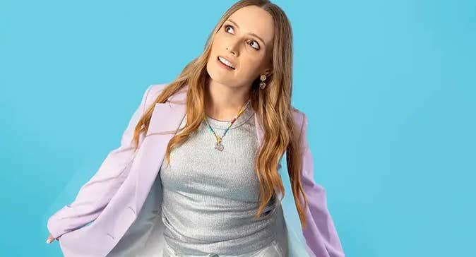 A woman is looking with interest up to her right, holding out either side of a lilac jacket against blue background.