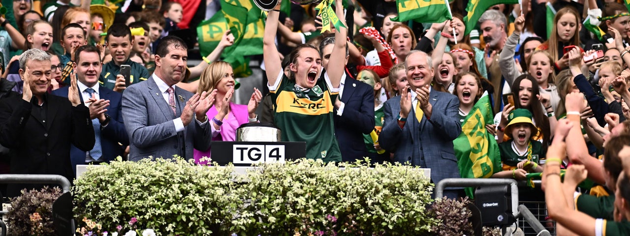 Celebrations in Croke Park, a sports person in jersey lifting large silver trophy over their head with clapping men either side in suits, against large crowd waving flags behind.