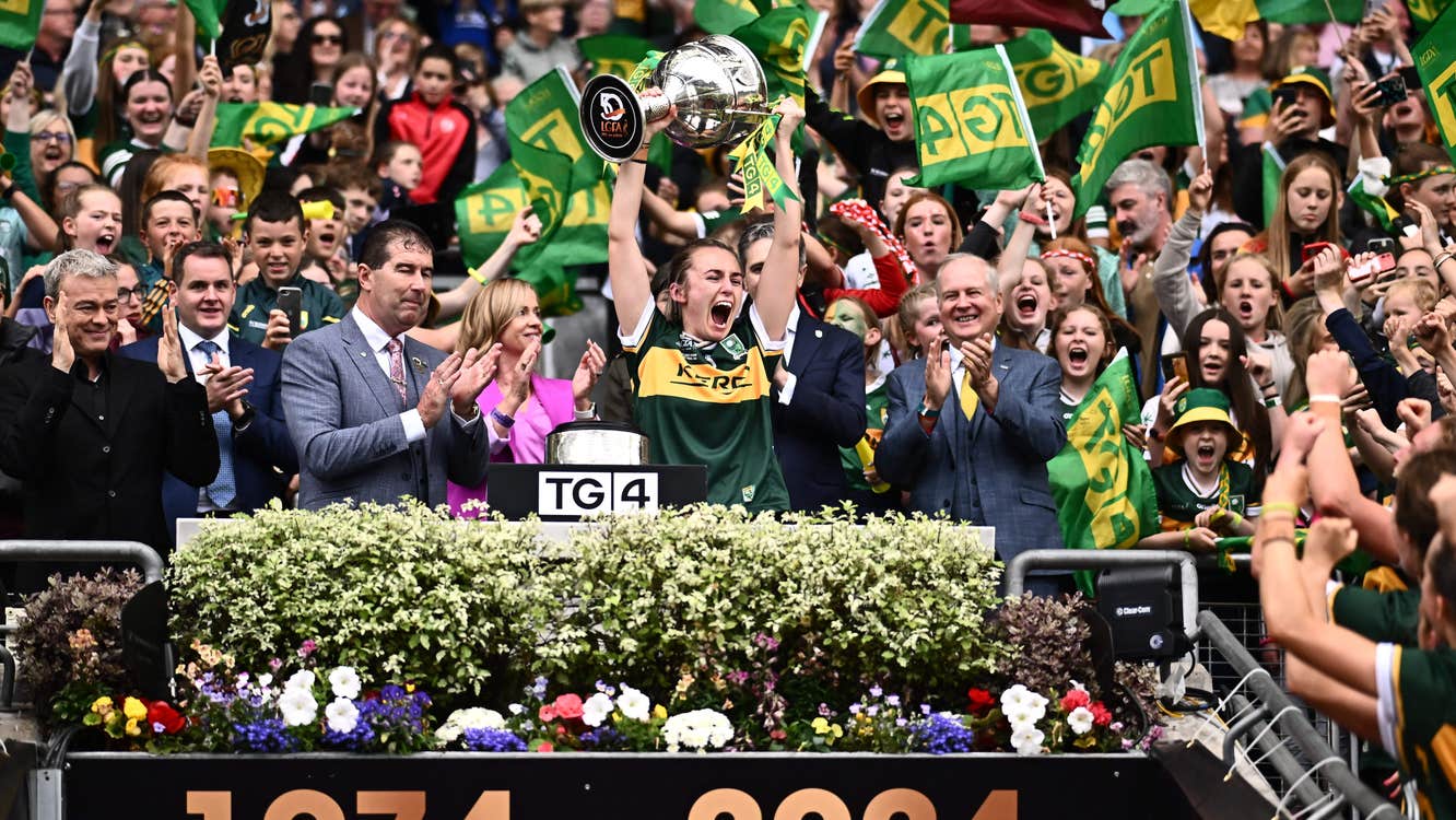 Celebrations in Croke Park, a sports person in jersey lifting large silver trophy over their head with clapping men either side in suits, against large crowd waving flags behind.