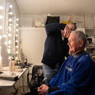 A man sitting in the make-up room of a theatre, in front of a mirror surrounded by lit light bulbs, having his hair done by another man.