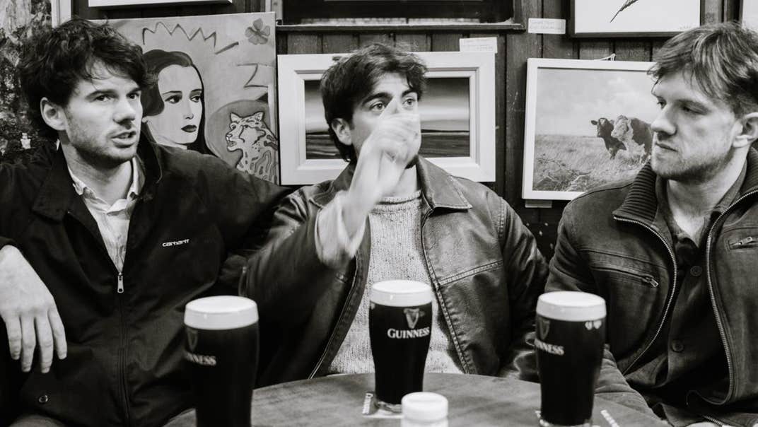 Black and white photo of 3 men sitting around a table with full pints of stout.