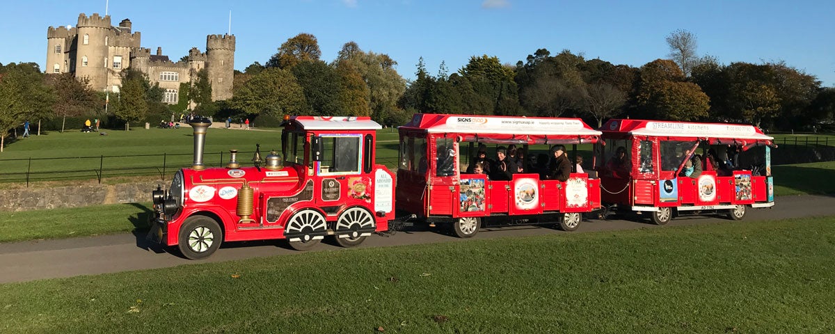 A small train running along a green park with a castle in the background