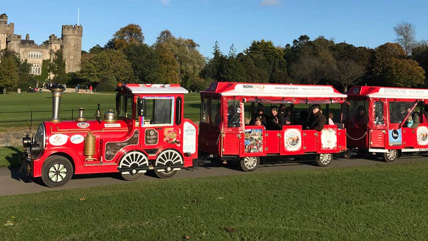 A small train running along a green park with a castle in the background
