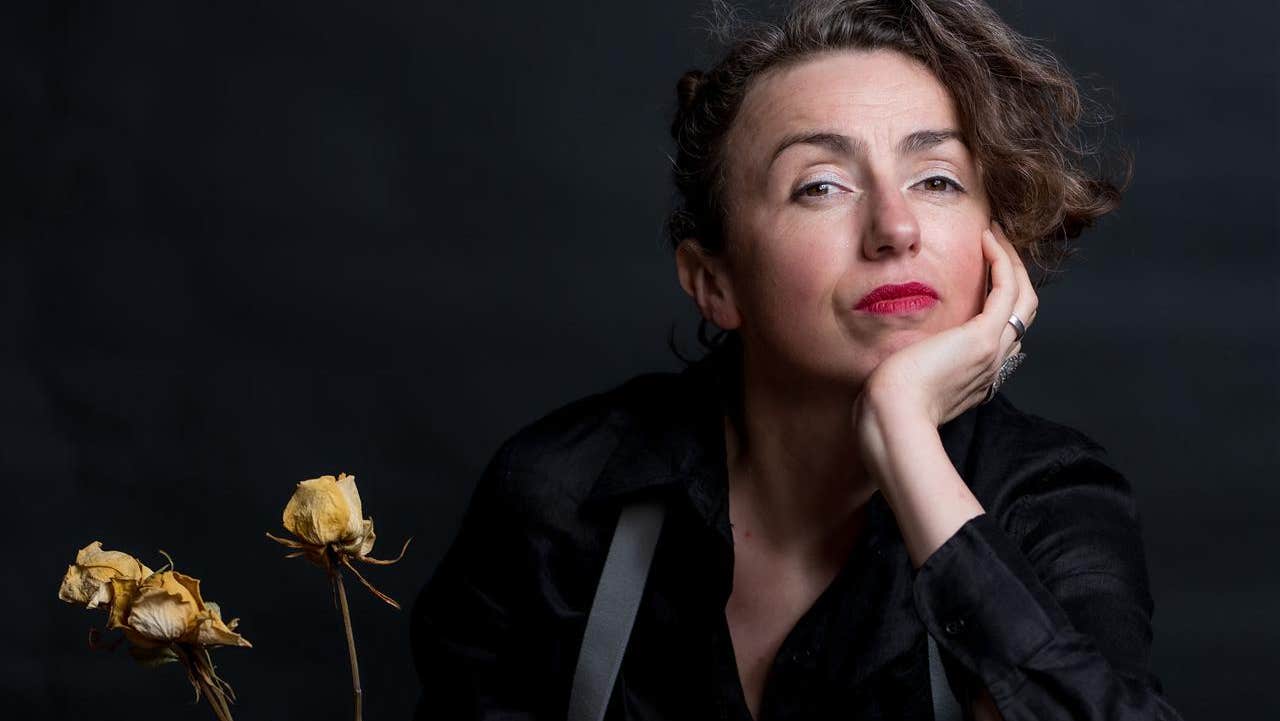 Against a black background Clare, wearing black clothes, grey suspenders and red lipstick with her left hand under her chin, her left foot on a chair, clasping a bottle and dead white roses.