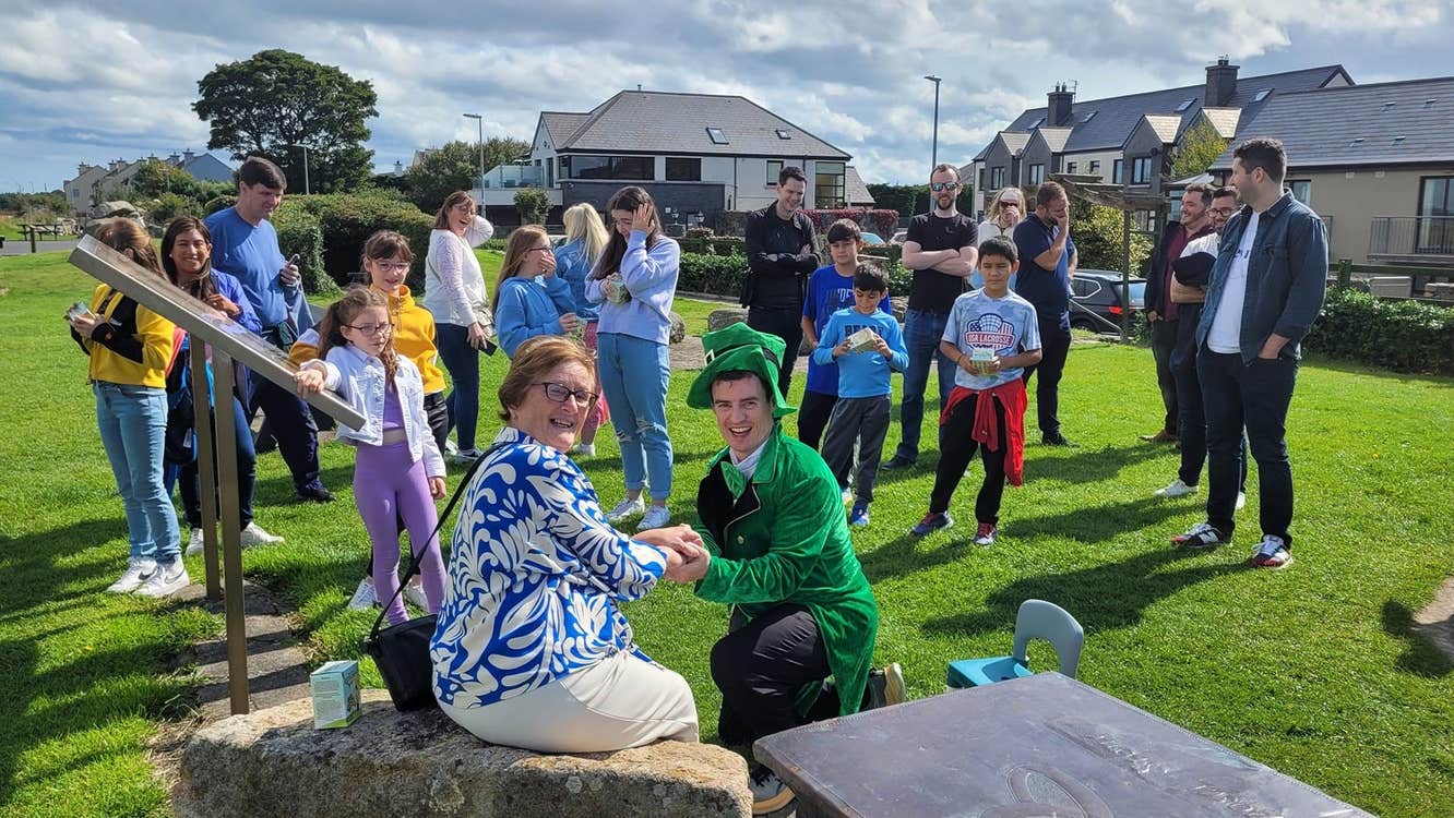 Lady sitting on a rock with a man in a leprechaun costume kneeling down and people in the background