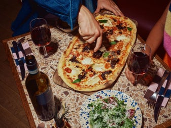 A close up of a pizza on a table with wine glasses as someone slices it