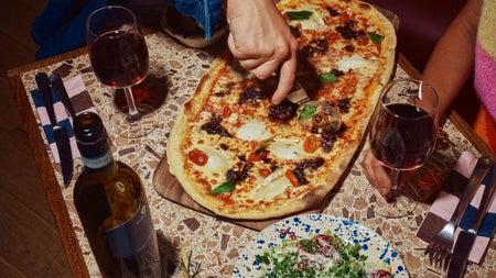 A close up of a pizza on a table with wine glasses as someone slices it