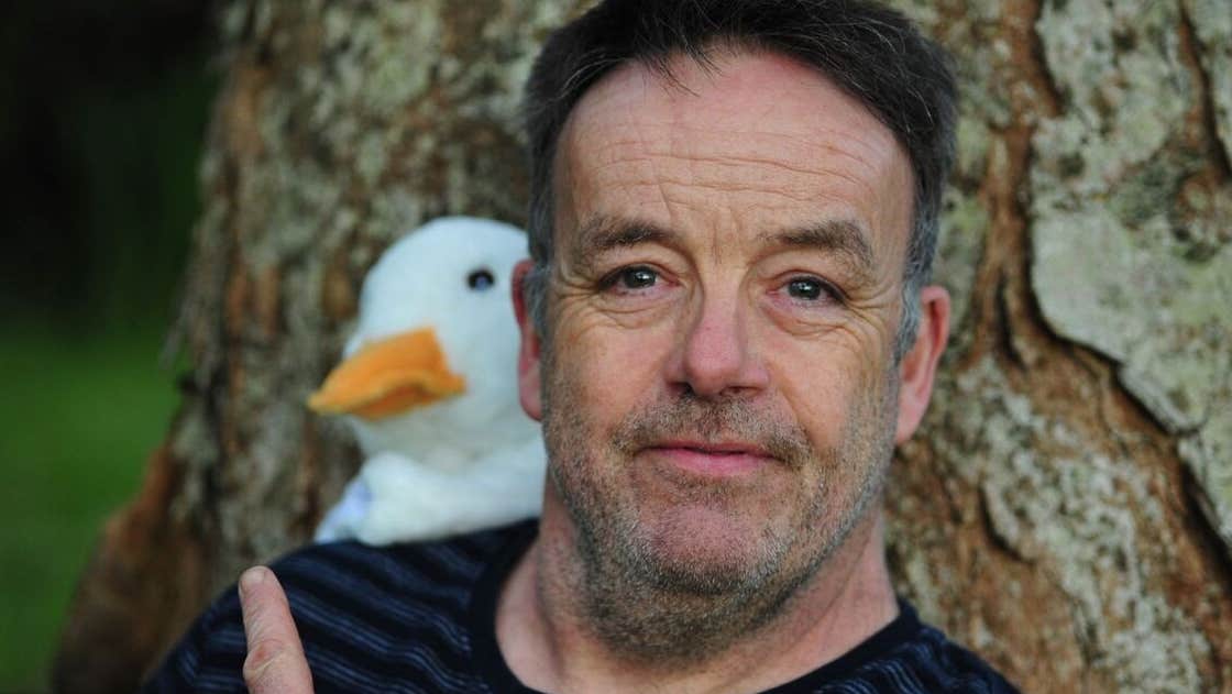 Púca – John Colleary: Mighty Quack, a close up of a man pointing over his shoulder at a white toy duck behind him