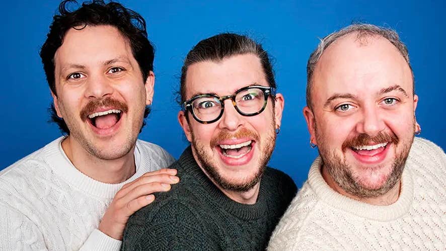 3 laughing men in a tight line wearing aran style knitted jumpers against plain blue background.