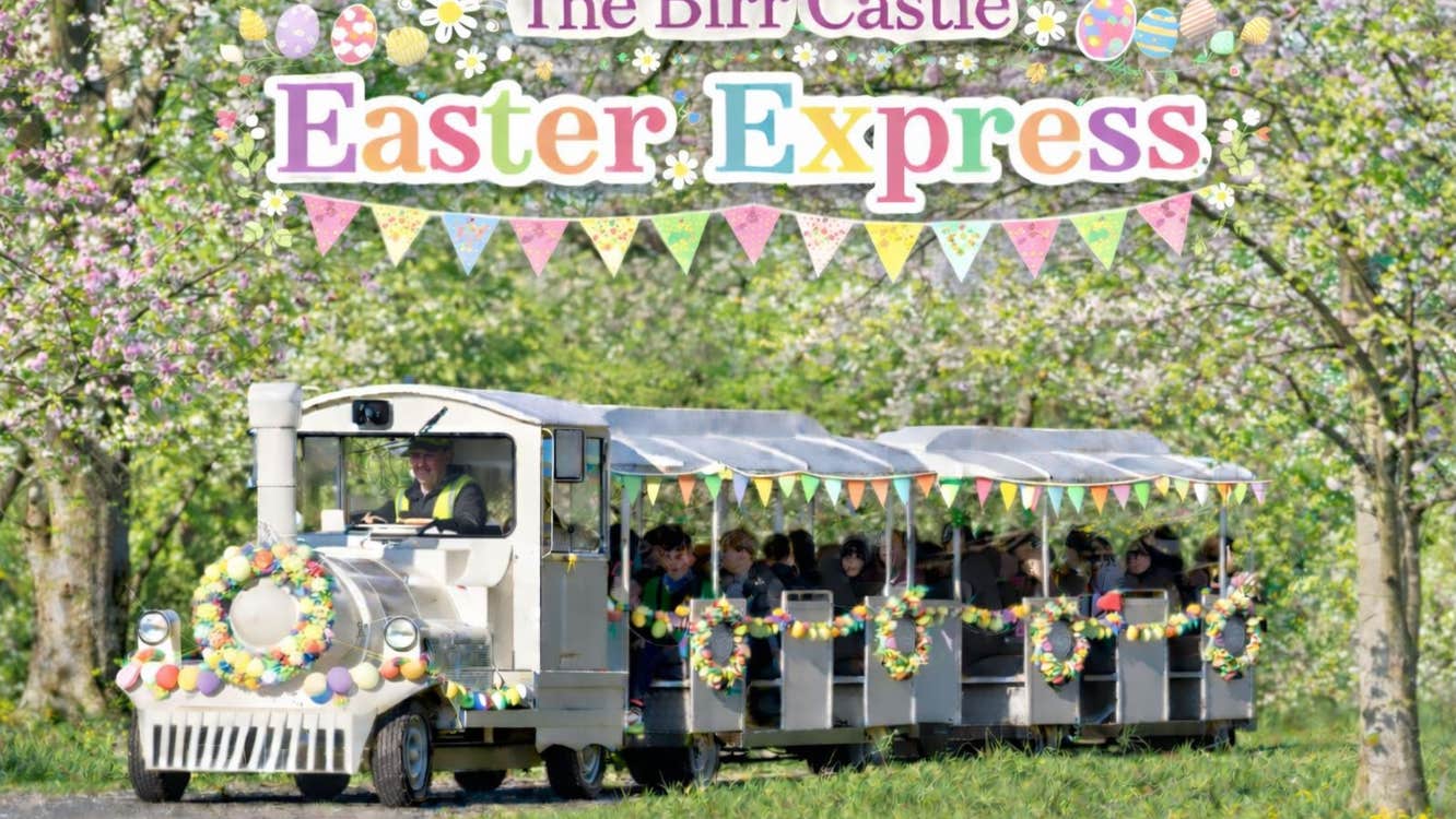 The Birr Castle Easter Express - a white train on a road pulling 2 decorated carriages in eggs and flowers in a woodland setting with blossom.