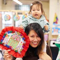 A smiling woman with a small child on her shoulders holding a craft made picture.