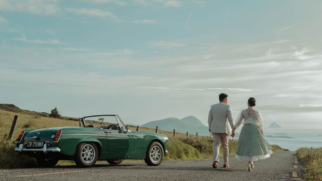 A couple walking hand in hand while passing a parked vintage car