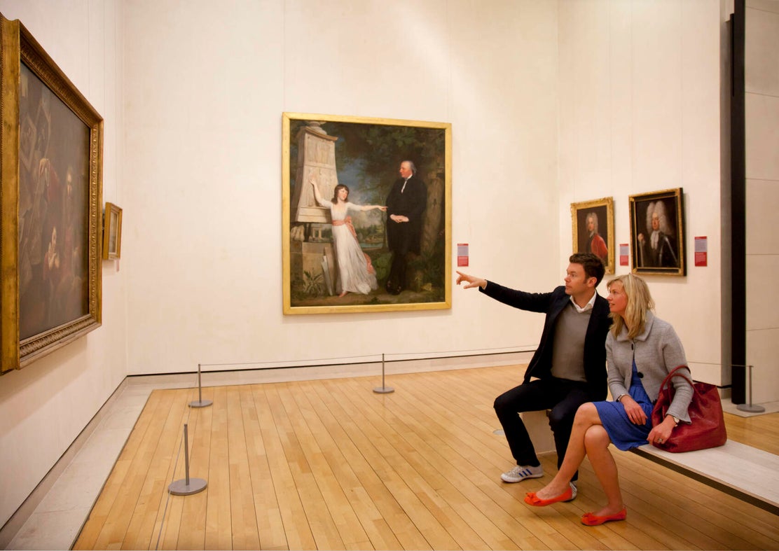 A couple sitting on a bench in the National Gallery of Ireland looking at a painting.