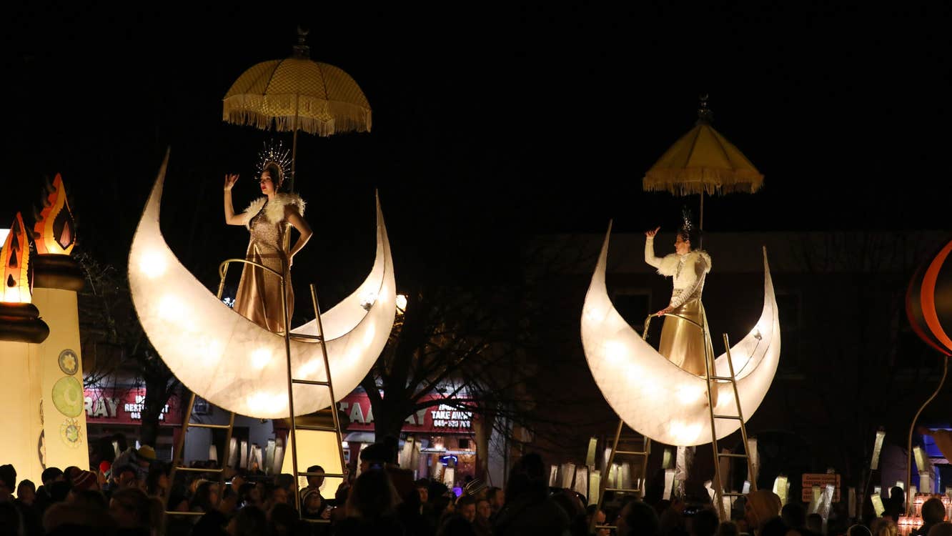 at night outdoors crowds are looking up at 2 illuminated half moon shapes each containing a person posing in a gold costume