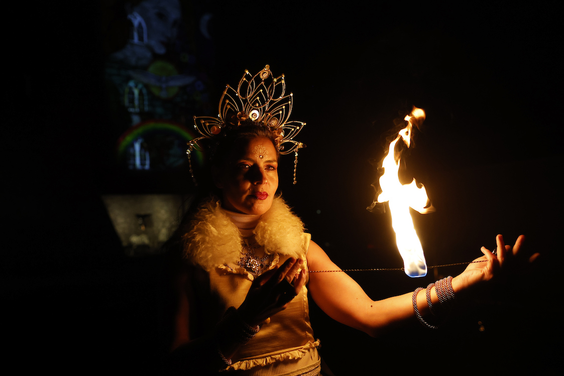 In the dark, a person with a decorative, wire headdress is holding a piece of thin wire between outstretched hands with fire burning in the middle.
