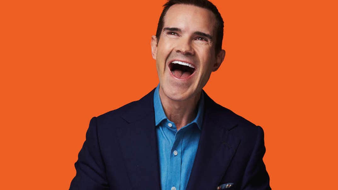 A man looking slightly up to his right is laughing with mouth open, in denim shirt and dark jacket, against bright orange background.