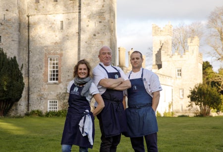 Three people in navy aprons posing on a green in front of a castle
