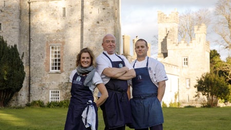Three people in navy aprons posing on a green in front of a castle