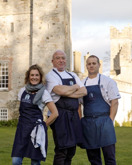 Three people in navy aprons posing on a green in front of a castle