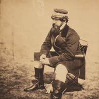 An old, brown style photo of a man in uniform with black boots seated on a camping chair.