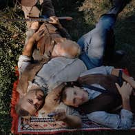 A photograph taken from standing height of three men dressed in Western style clothing lying on a woven rug on a grassy terrain. The men are of various ages, each resting his head upon the other.