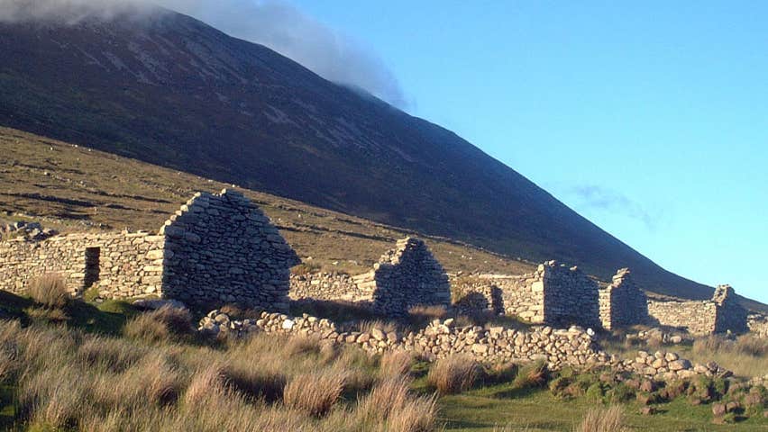 The ruins of stone cottages at the foot of a mountain with mist rolling down from the top of the mountain