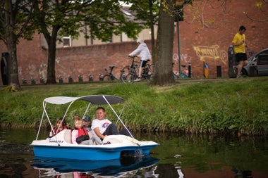 Man and three children in a blue and white pedal boat sailing down a canal with a grass bank to the right