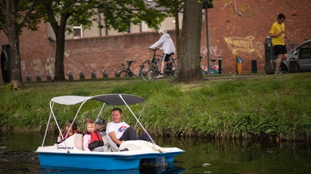 Man and three children in a blue and white pedal boat sailing down a canal with a grass bank to the right
