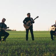 3 men with guitars in a grassy field, 2 seated on chairs.