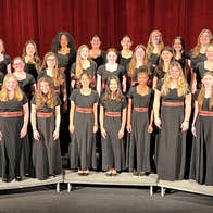 Sacred Heart Madrigals and Upper School Chorus - a group of young women standing on 3 levels, all smiling, wearing long, dark dresses with red band around the middle.