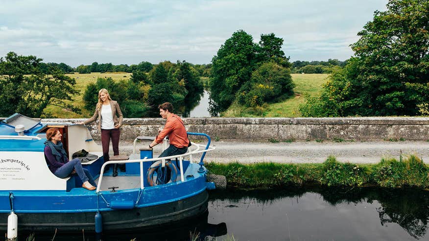 Three people onboard a barge with a backdrop of green fields and trees