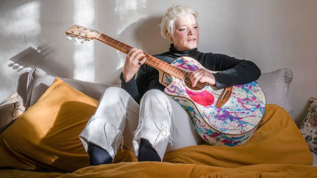A woman seated on a bed playing a painted splattered guitar.
