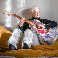 A woman seated on a bed playing a painted splattered guitar.