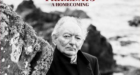 Black and white image of an older man standing beside a rock at the water's edge.