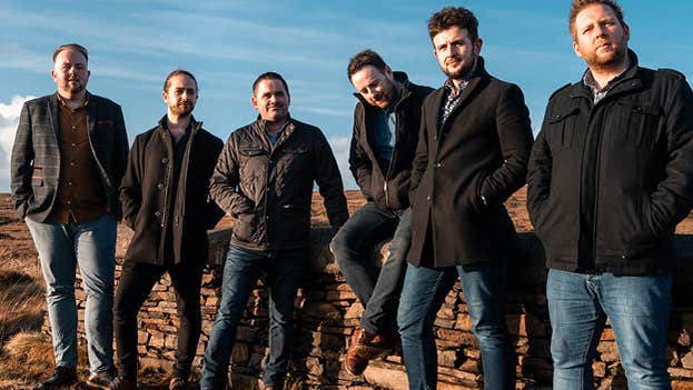 A group of 6 men in coats are standing in a line on the edge of a road with bogland behind.
