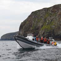 People on a grey rib boat in the water wearing orange life jackets in front of cliffs