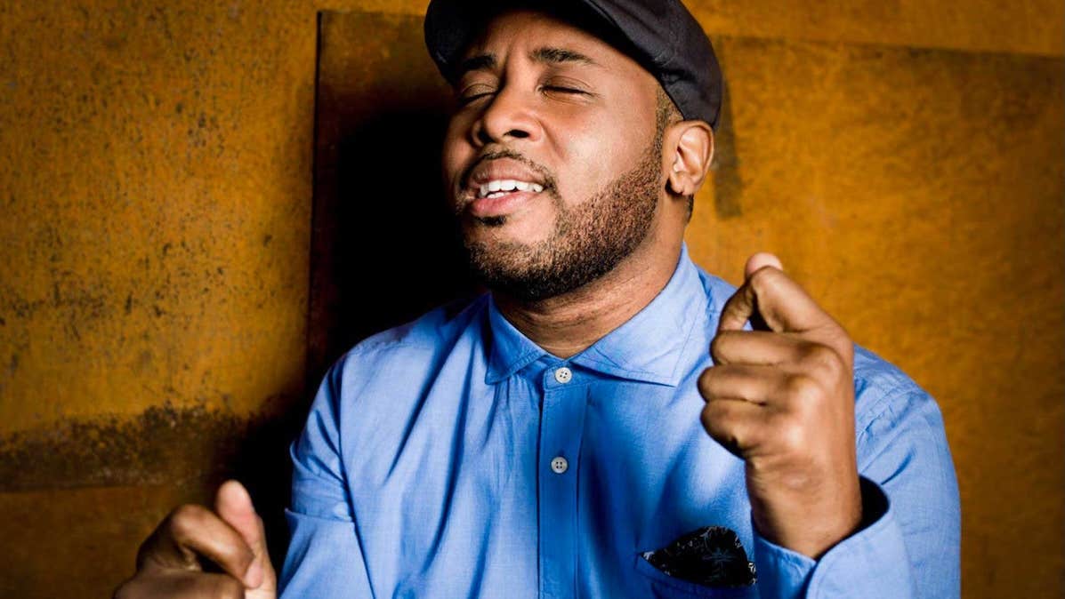 A person singing with eyes closed holding hands up in fists, standing beside a brown wall