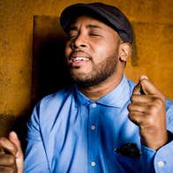 A person singing with eyes closed holding hands up in fists, standing beside a brown wall