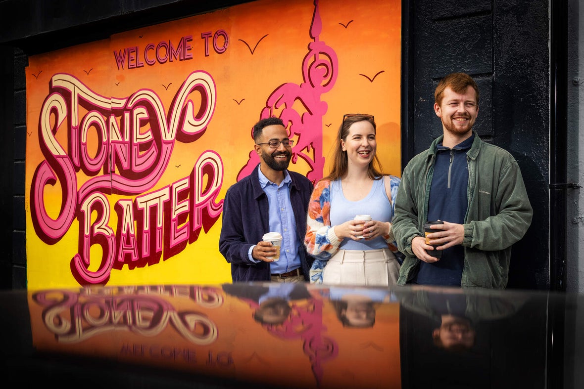 A few friends holding coffee and laughing at something off camera next to the Welcome to Stoneybatter sign.