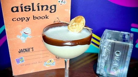 A creamy cocktail in a coupe glass beside an aisling copy book and cassette tapes