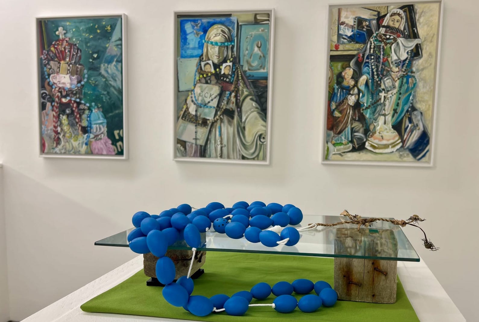 3 paintings on a gallery wall with a table in front with a string of large blue beads on a glass display plinth.