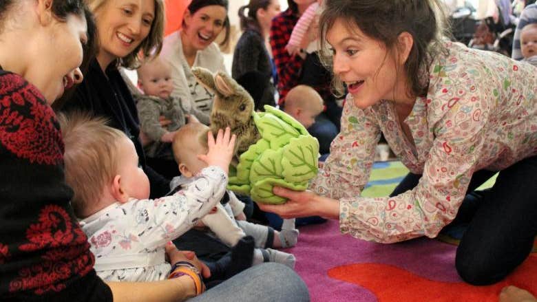 A woman is smiling and holding up a soft toy of a rabbit in a lettuce to a small baby on a parent's lap with others looking on.
