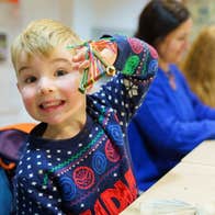 A smiling young boy in a Christmas jumper seated at a table holding up a small handmade decoration