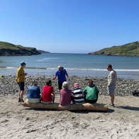 A group of people on a hiking tour sit on a log on a beach listening to their tour guide