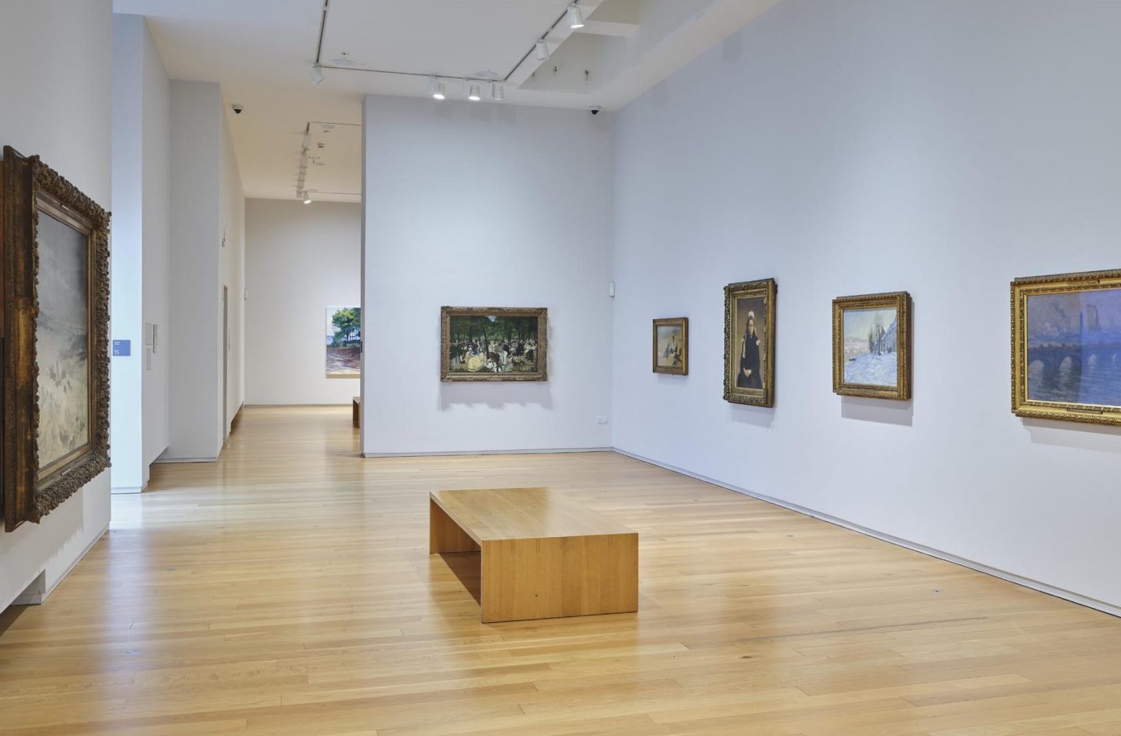 View of a gallery room with pale walls and light wood floor and small wooden bench in the centre, different size art works along each wall.