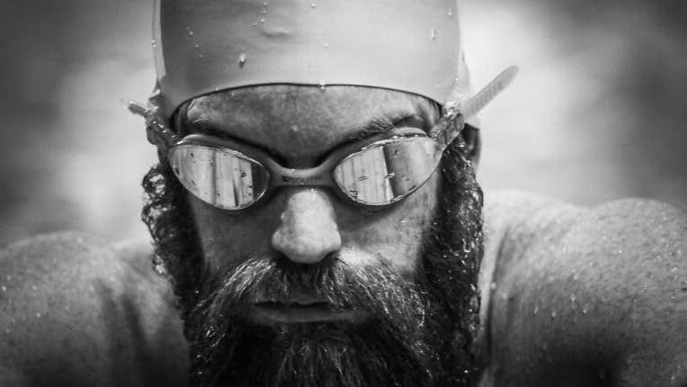 Black and white headshot of man with a beard wearing swim goggles and hat covered in water droplets.